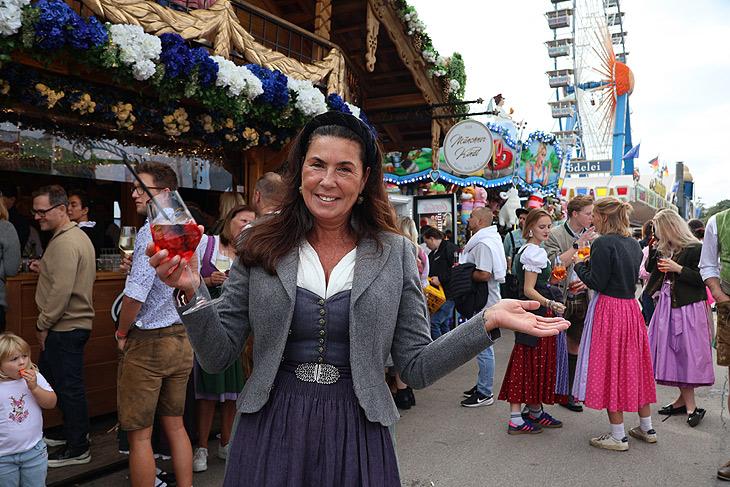 Miriam Blume steht mit dem "Zum Münchner Kindl" Sekt, Wein & Co. 2025 auf der Strasse 5 mittig zwischen Olympialooping und Riesenrad (©Foto: Martin Schmitz) 